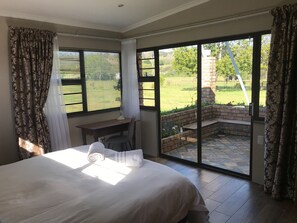 A bedroom with a large bed, a desk, and a view of an outdoor patio with a brick fireplace.