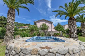 A stone pool with a hot tub in a garden with palm trees and a pink building in the background. - Apartment 'Bilocale Dolores' with Shared Pool, Wi-Fi and Air Conditioning (Campo nell'Elba)