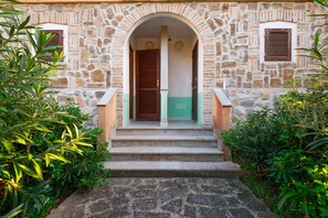 Un bâtiment en pierre avec une porte d’entrée centrale, flanquée de deux fenêtres avec volets, et un escalier menant à l’entrée.