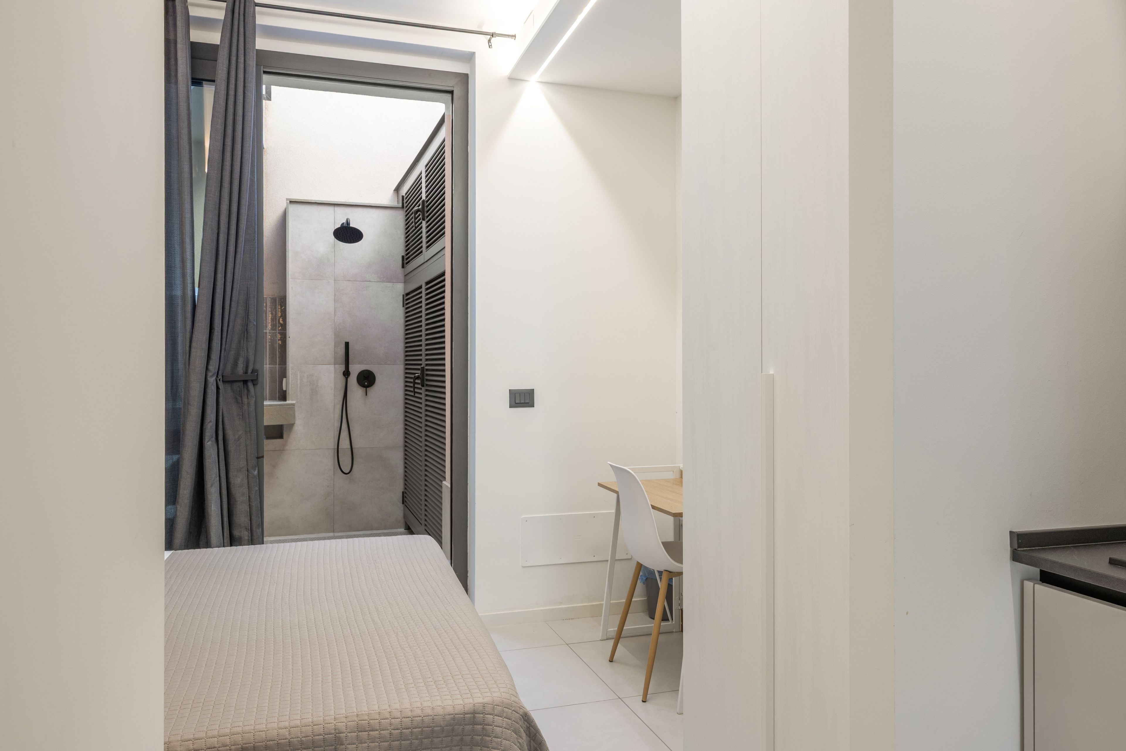 Een smalle gang die leidt naar een badkamer met een douche, een bed en een klein tafeltje met een stoel.