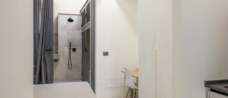 A narrow hallway leading to a bathroom with a shower, a bed, and a small table with a chair.