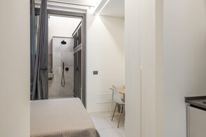 Een smalle gang die leidt naar een badkamer met een douche, een bed en een klein tafeltje met een stoel.