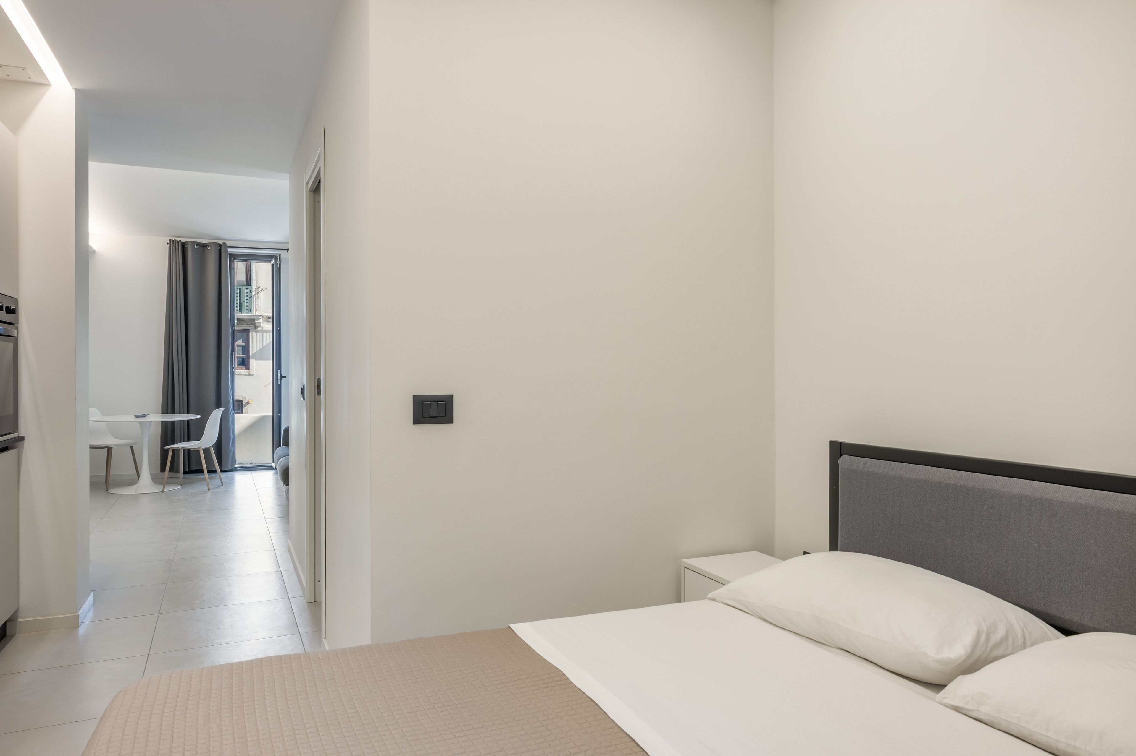 A hotel room with a bed, bedside tables, a desk with chairs, and a view through an open door to a balcony.