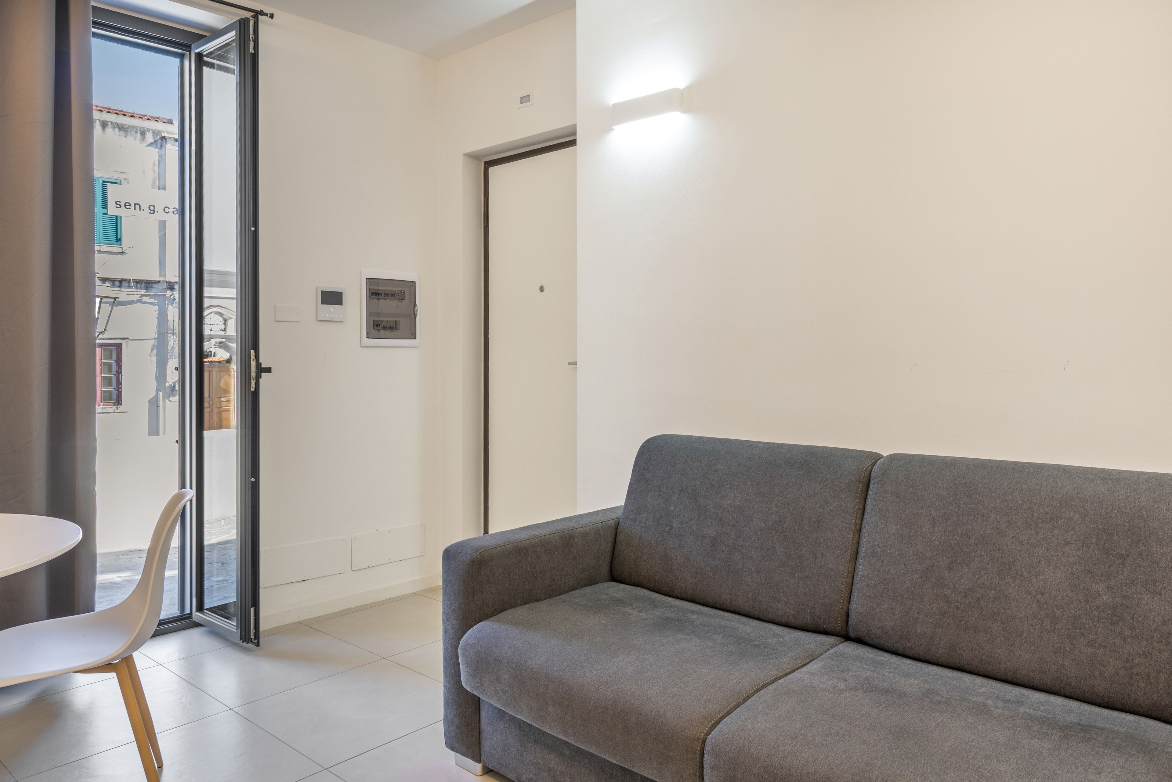 Een modern interieur met een grijze bank, een witte tafel en een glazen deur die leidt naar een buitenruimte.