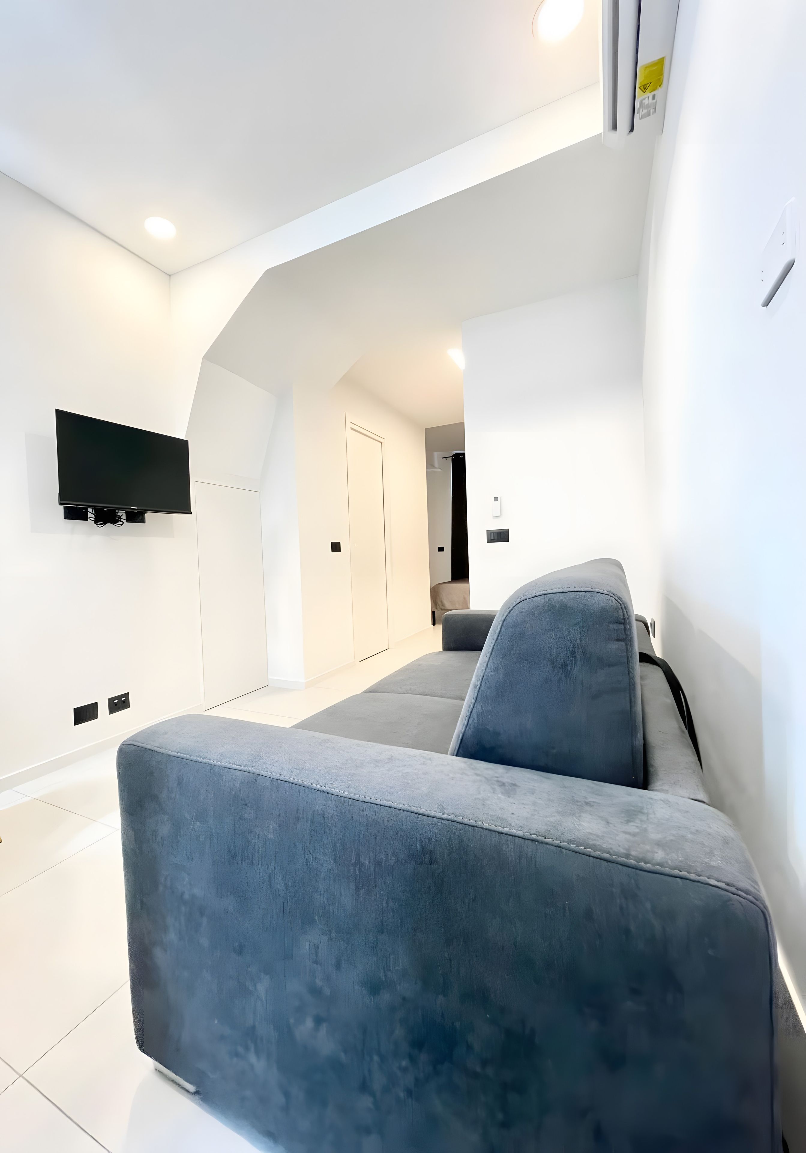 Una sala de estar moderna con un sofá gris, un televisor de pantalla plana montado en la pared y una puerta que da a otra habitación.