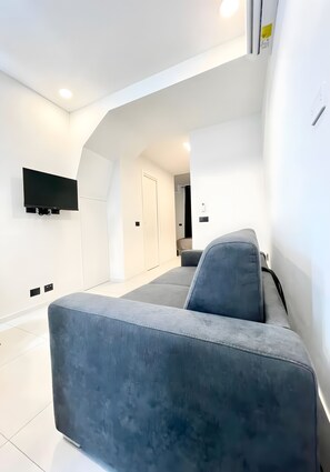 A modern living room with a grey sofa, a flat-screen TV mounted on the wall, and a door leading to another room.