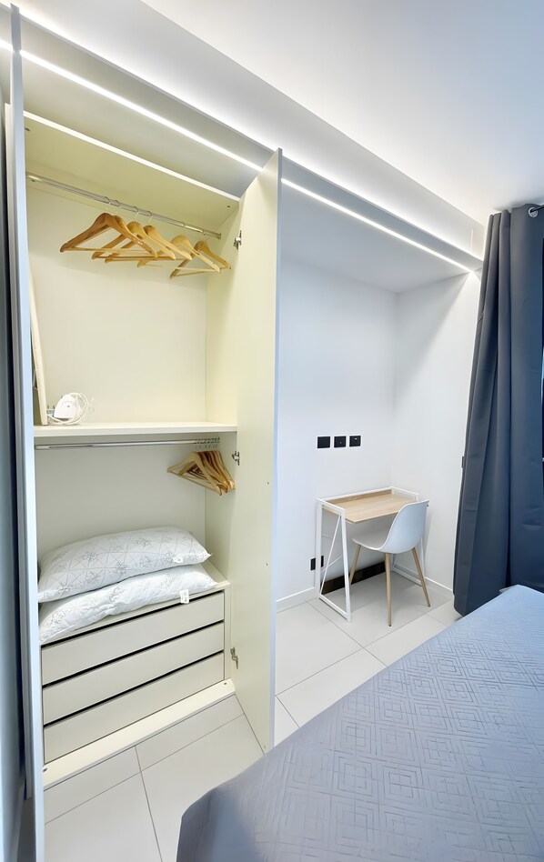 A modern bedroom with a bed, a desk, and a wardrobe with hanging clothes.