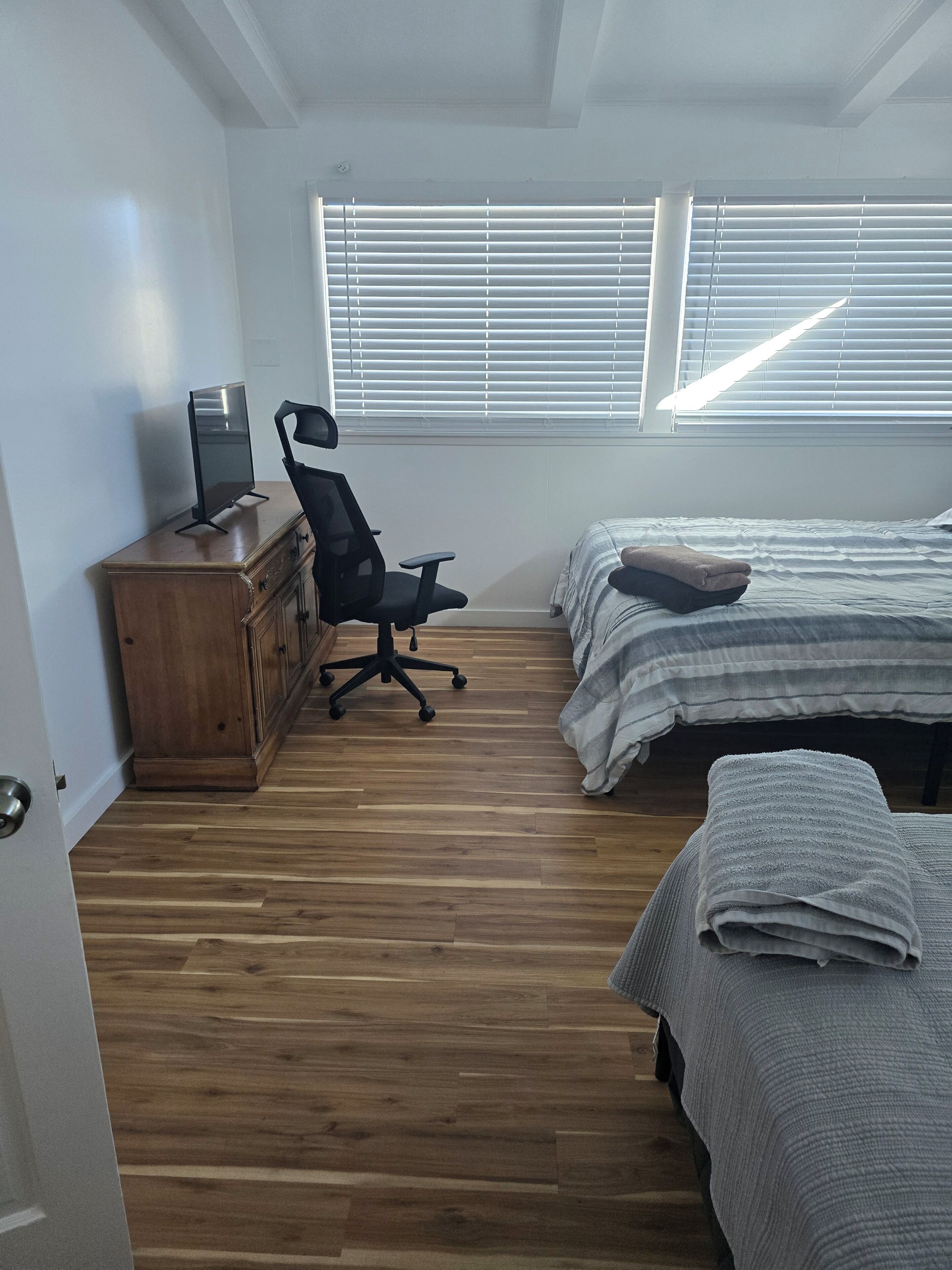 Une chambre avec un plancher en bois, un bureau avec une chaise, un lit avec une couverture et une fenêtre avec des stores.