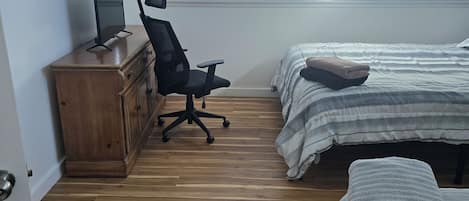 Une chambre avec un plancher en bois, un bureau avec une chaise, un lit avec une couverture et une fenêtre avec des stores.