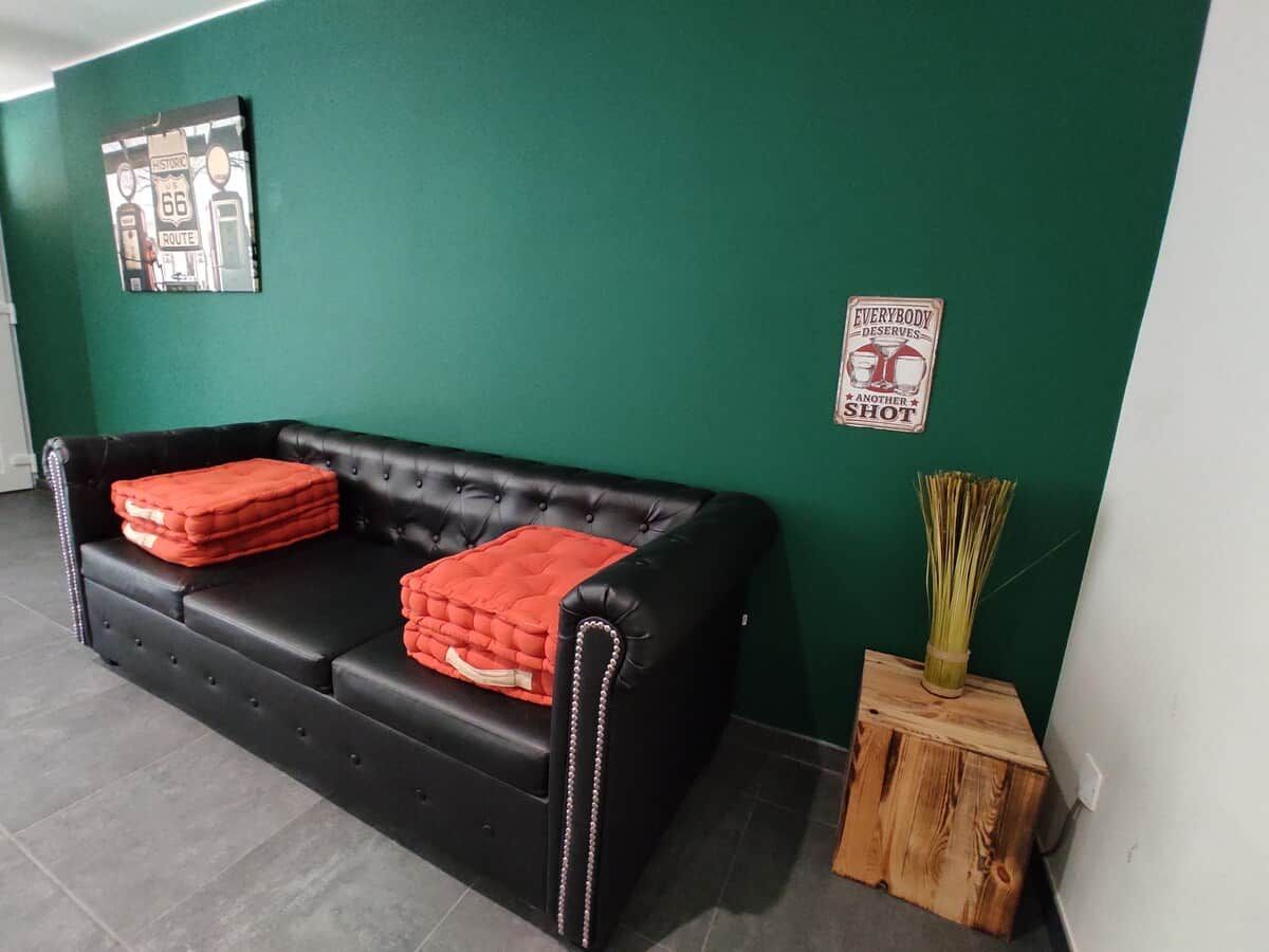Een zwarte leren bank met rode kussens, een groene wand, een klein houten bijzettafeltje met een vaas, en een poster aan de muur.