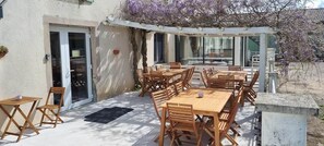 Een terras buiten met houten tafels en stoelen, een overdekte zithoek en uitzicht op een gebouw met een schuifdeur.