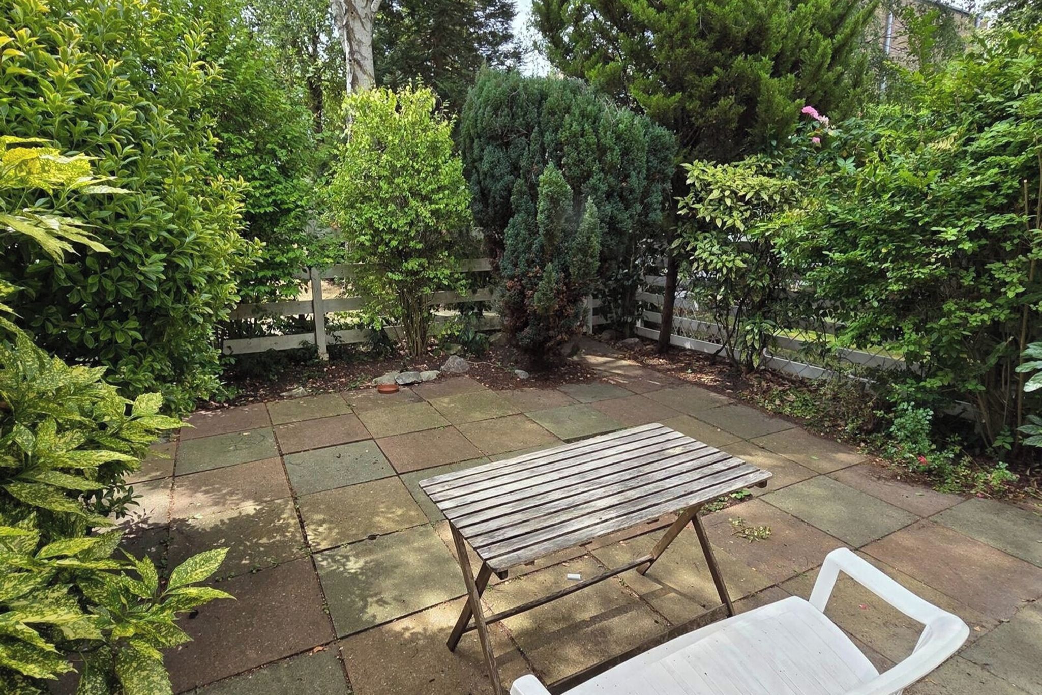 Une terrasse avec une table et des chaises en bois, entourée d’une végétation luxuriante et d’une clôture en bois.