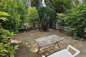 Un patio con una mesa y sillas de madera, rodeado de abundante follaje y una valla de madera.
