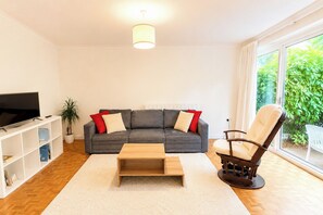 A living room with a grey sofa, a wooden coffee table, a rocking chair, a TV on a white stand, and a bookshelf. - Bright 2BDR Flat with King Bed in West London (Greater London)