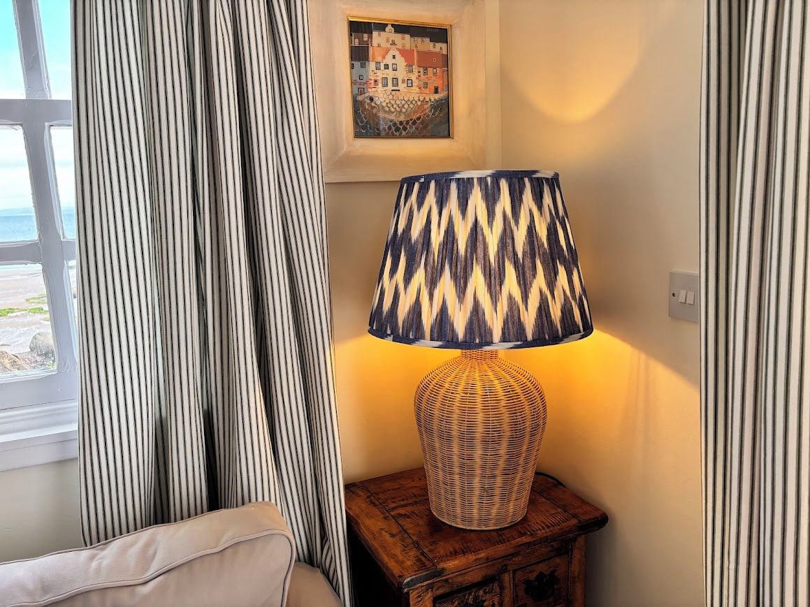 A room with a lamp on a wooden nightstand, a window with curtains, and a framed picture on the wall.