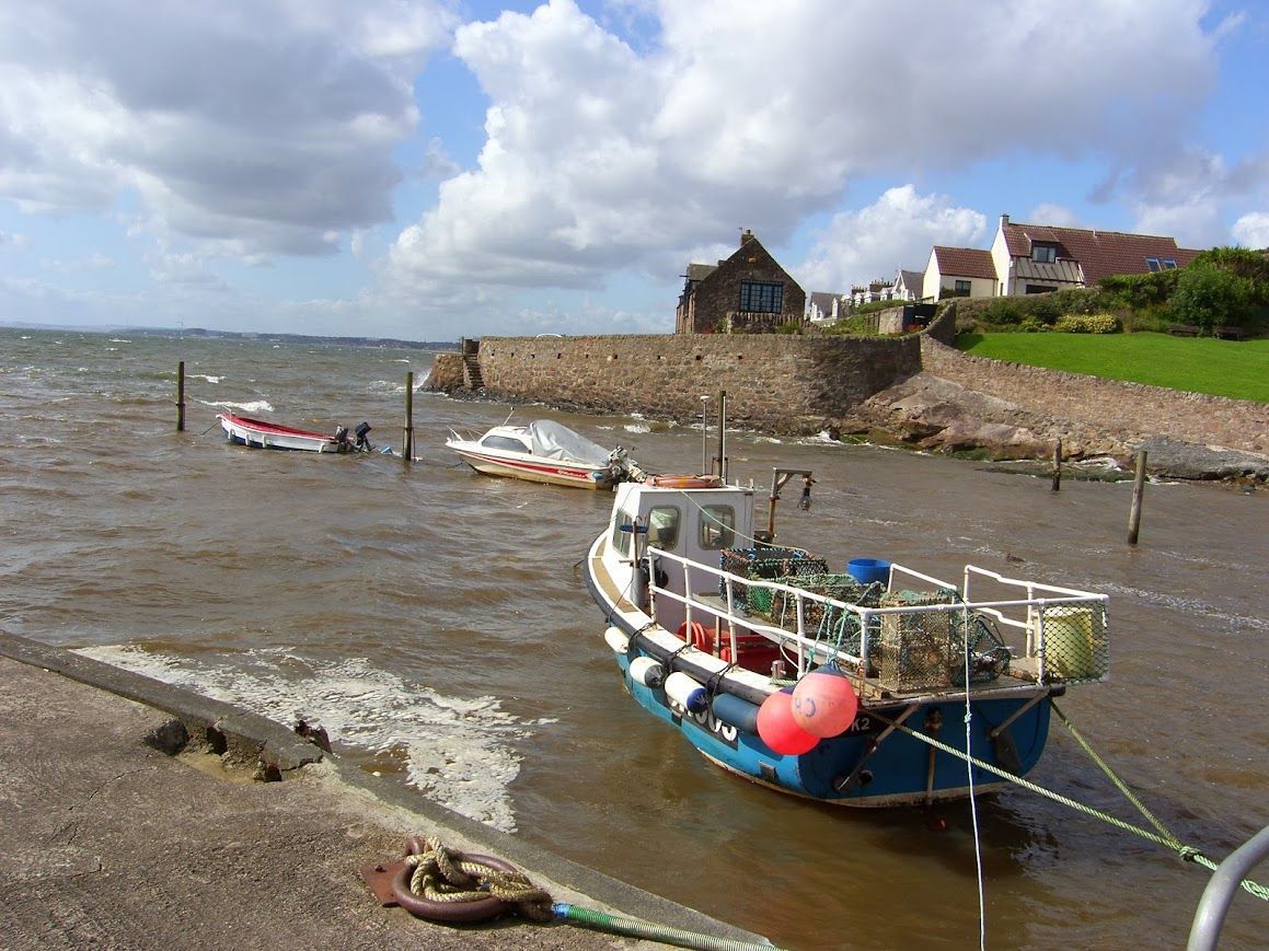 A coastal scene with a boat docked at a stone pier, a house with a red roof, and a grassy area.