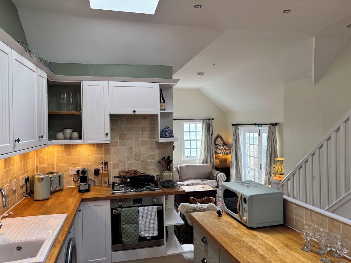 A compact kitchen with a built-in oven, a microwave, and a toaster. There is a window with curtains, a wooden countertop, and a staircase leading to another level.