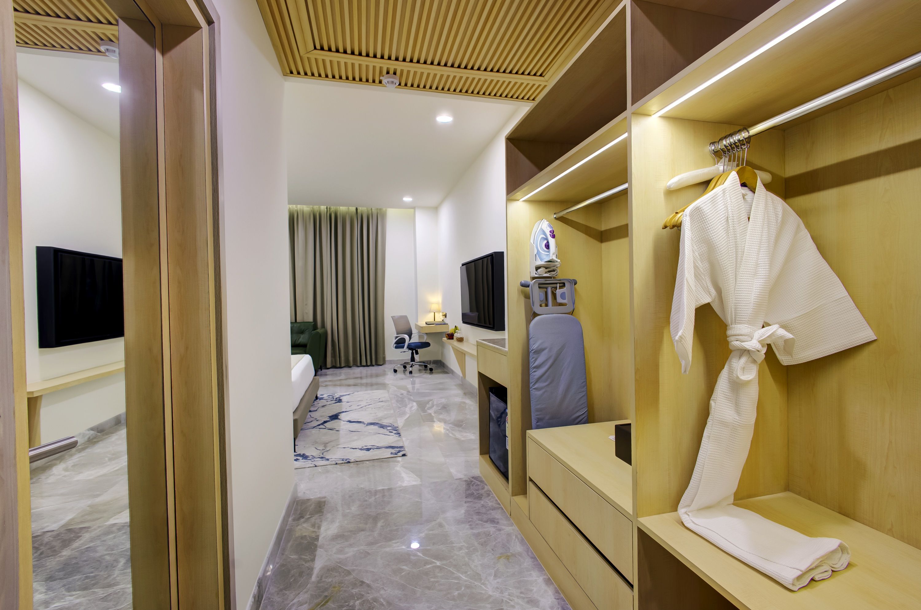 A hotel room with a wooden wardrobe, a flat-screen TV, a desk with a chair, a robe hanging on the wardrobe, and a bed with a green headboard.