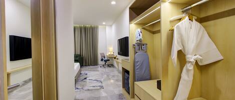 A hotel room with a wooden wardrobe, a flat-screen TV, a desk with a chair, a robe hanging on the wardrobe, and a bed with a green headboard.