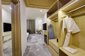 A hotel room with a wooden wardrobe, a flat-screen TV, a desk with a chair, a robe hanging on the wardrobe, and a bed with a green headboard. - Cygnett Collection (Gurugram)