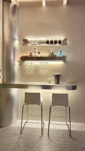 Un área de bar con dos sillas, un estante con botellas y vasos, y una repisa iluminada arriba.