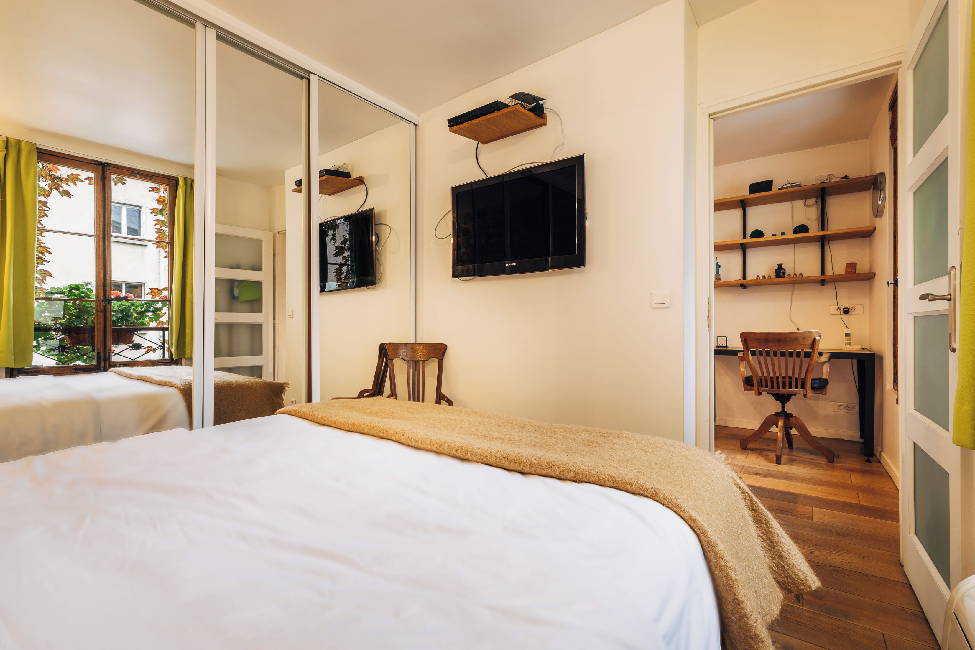 A bedroom with a bed, a television mounted on the wall, a desk with a chair, and a sliding door leading to a balcony.