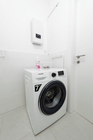 A compact, white washing machine with a digital control panel, a small bottle of fabric softener, and a white door in a clean, minimalistic bathroom. - Ardell - Aceessible Location - Comfortable 1BR - FREE Garage (Vienna)