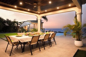 Een terras met een eettafel voor zes personen, omringd door stoelen, met uitzicht op de oceaan en een prieeltje op de achtergrond.