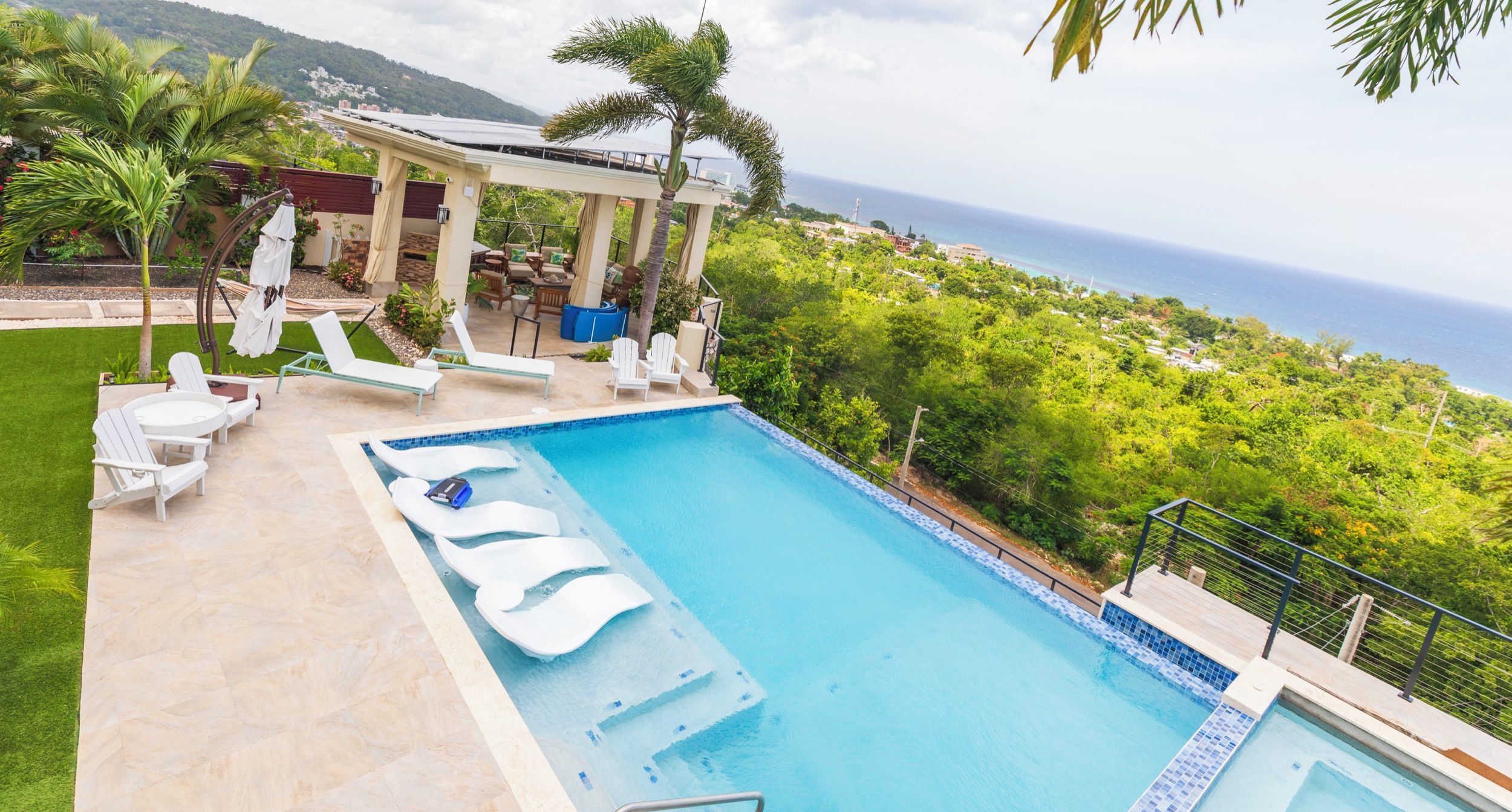A swimming pool with lounge chairs and a covered seating area, surrounded by greenery and overlooking a coastal landscape.