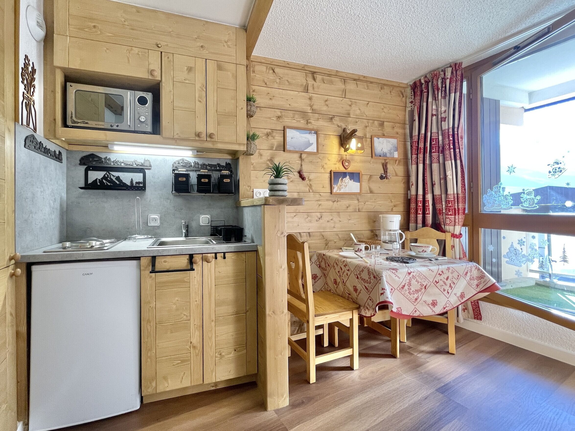 Eine kompakte Küche mit Holdekor-Schränken, einer Mikrowelle, einem Spülbecken und einem kleinen Kühlschrank. Ein Esstisch mit Stühlen und Tischdecke lädt zum Verweilen ein, und ein Fenster mit Vorhängen sorgt für angenehme Lichtverhältnisse.
