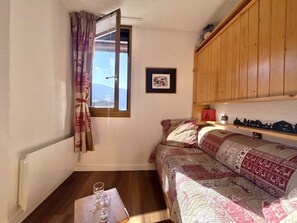 A cozy room with a bed, a wooden shelf with decorative items, a framed picture on the wall, and a window with curtains. - (SO.317) Alpine-style studio, right at the slopes, Les (Les Menuires)