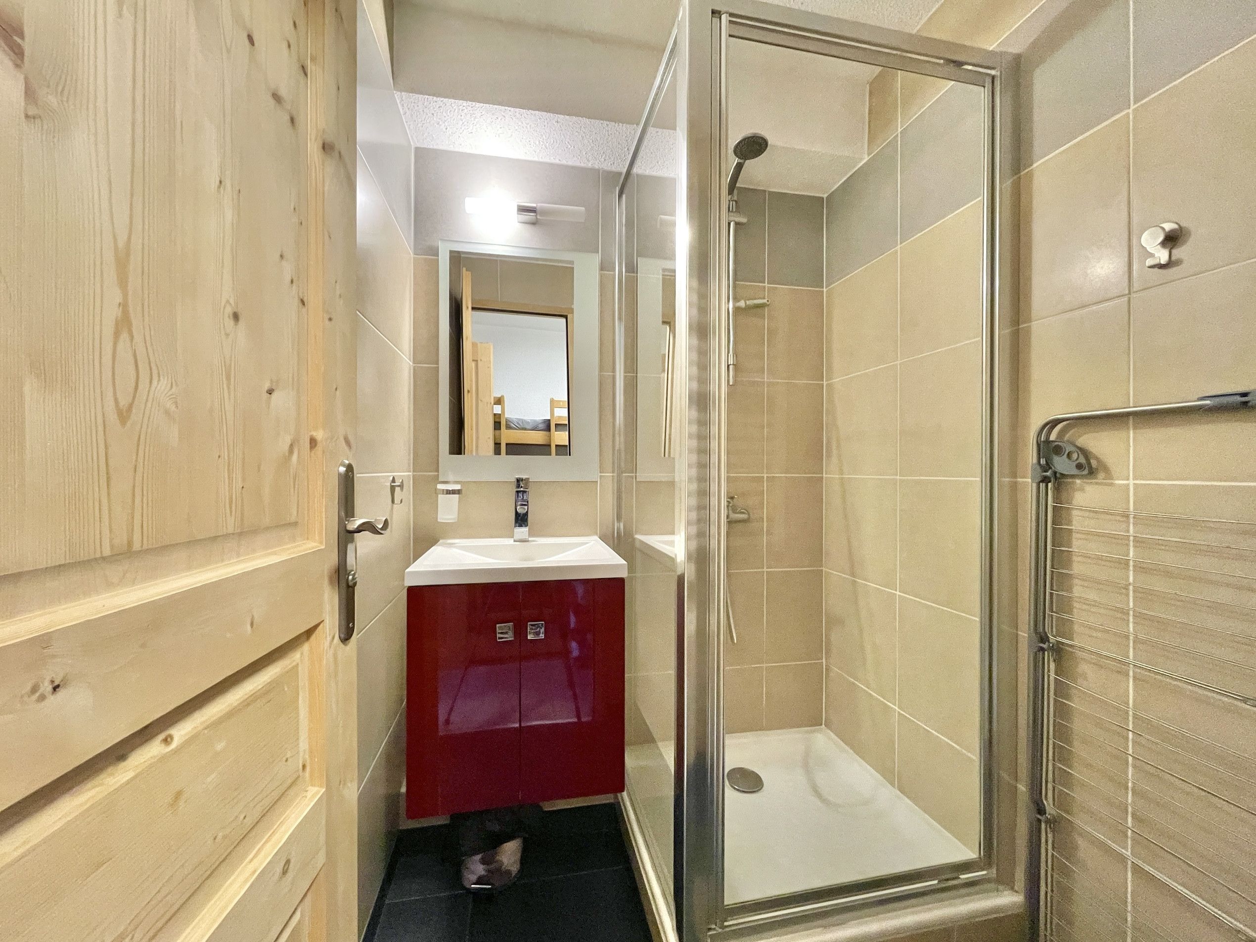 Une salle de bain avec une porte en bois, un meuble rouge, un miroir, une douche et un espace de douche clos par des parois de verre.
