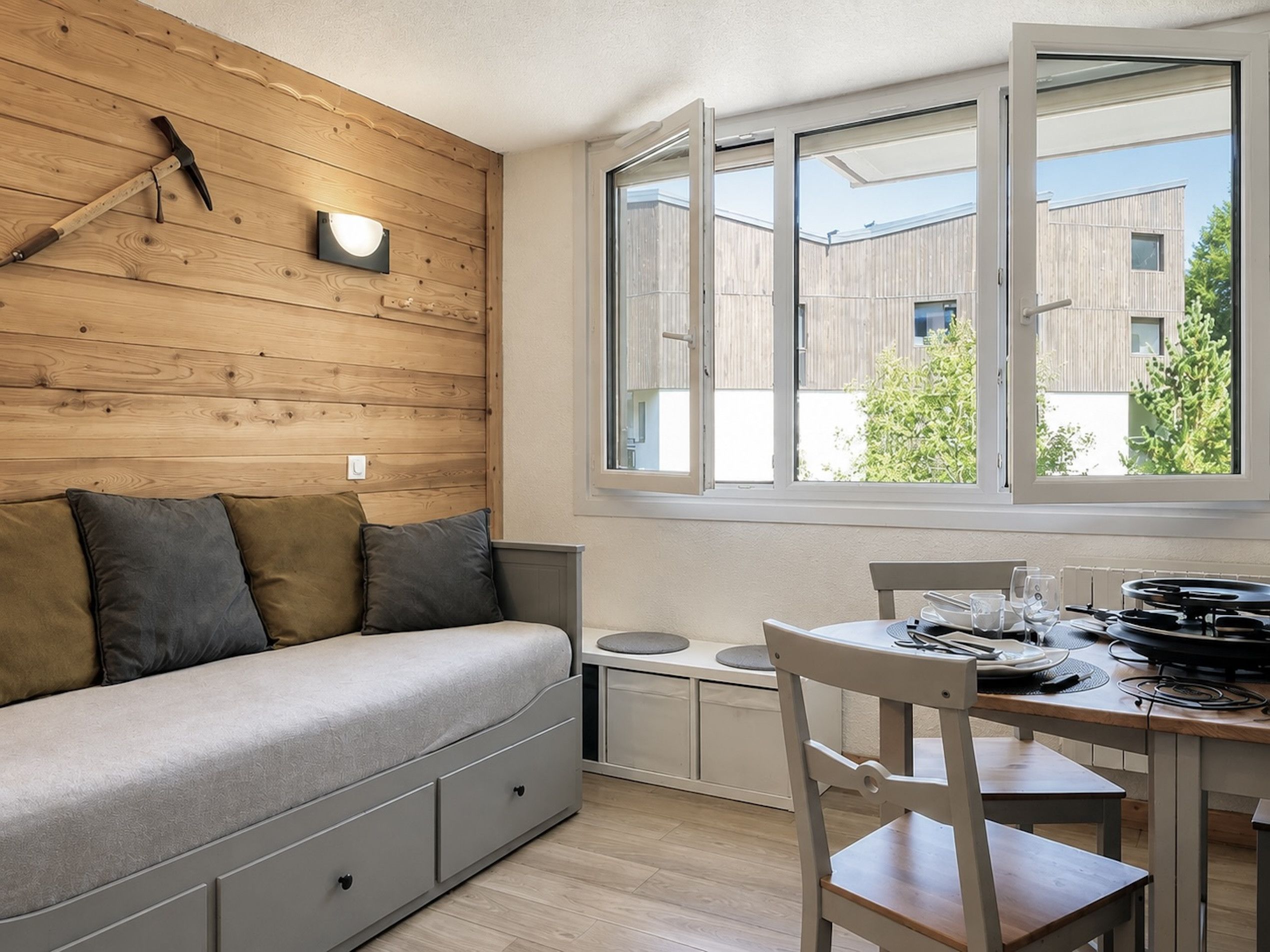 A modern room with a wooden wall, a sofa, a dining table with chairs, and a window with a view of buildings and trees.