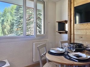 A dining area with a table set for two, a chair, a radiator, and a window with a view of trees. - (BO.421) Comfortable studio apartment, right on the slopes in 3 Vallées (Les Menuires)