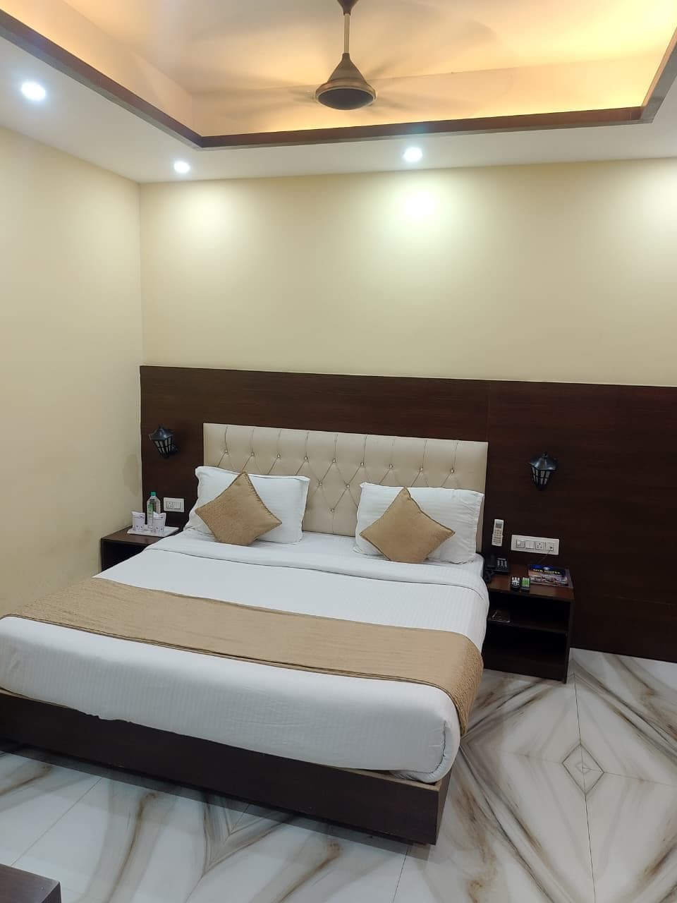 Una habitación de hotel con una cama, mesitas de noche, un ventilador de techo y suelo de mármol.