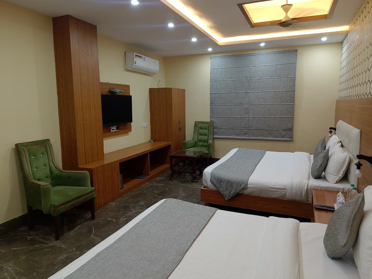 Habitación de hotel con dos camas, un televisor, una silla verde, un armario de madera y una pared gris con ventana.