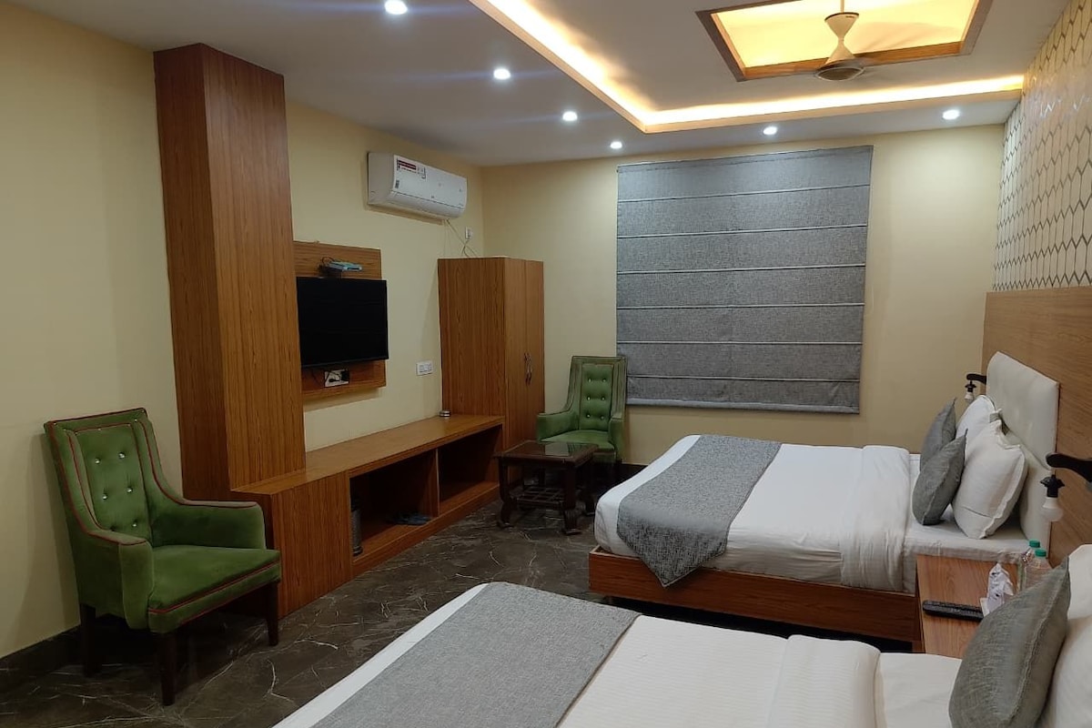 Habitación de hotel con dos camas, un televisor, una silla verde, un armario de madera y una pared gris con ventana.