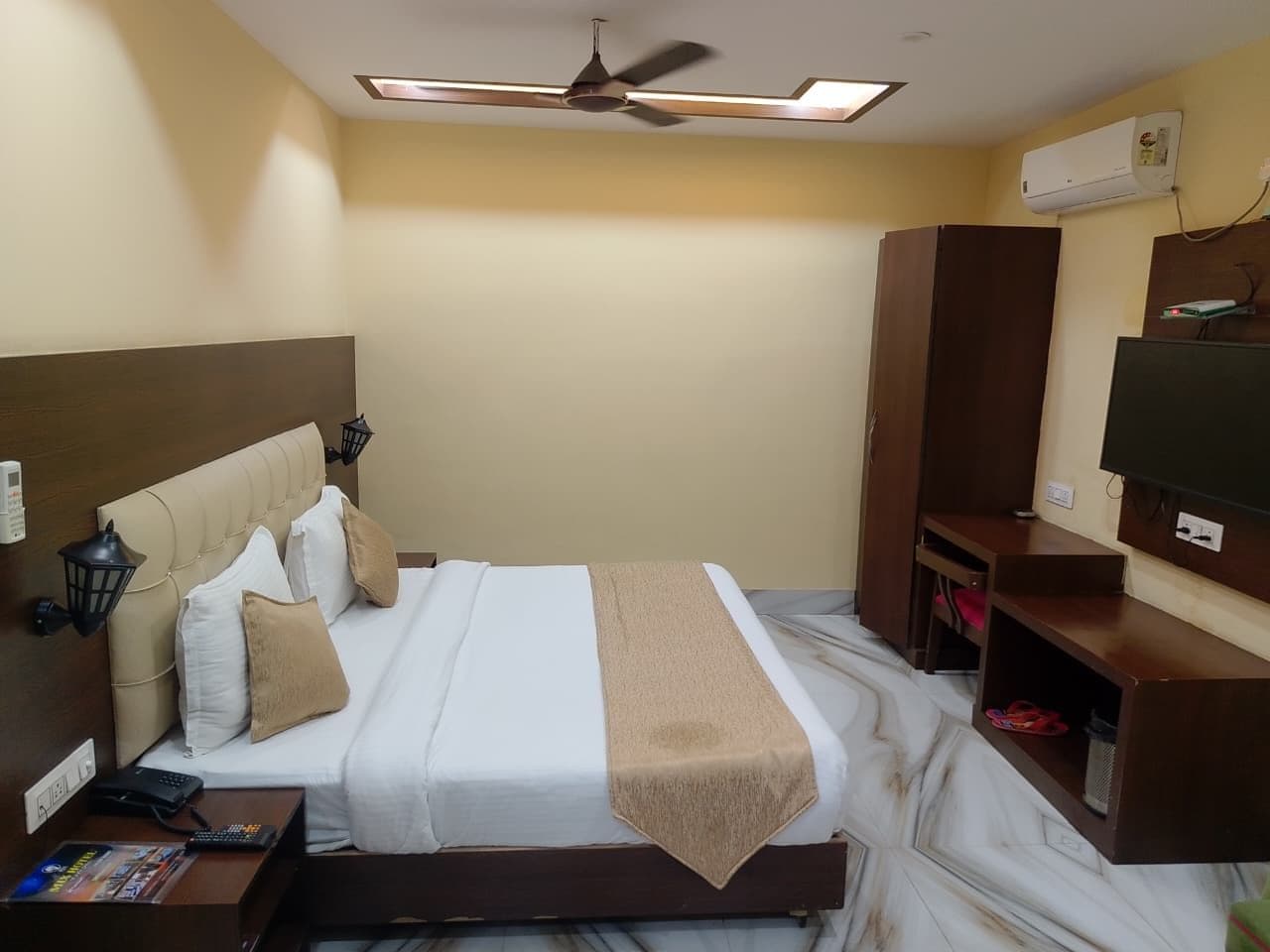 A hotel room with a bed, a ceiling fan, an air conditioning unit, a TV, and a desk with a telephone.