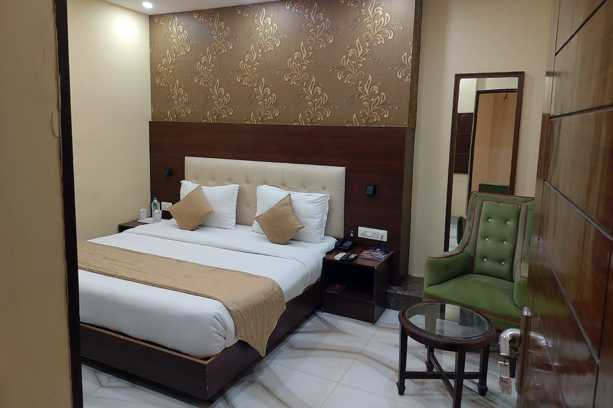 Una habitación de hotel con una cama grande, un sillón verde, una mesita y un espejo.