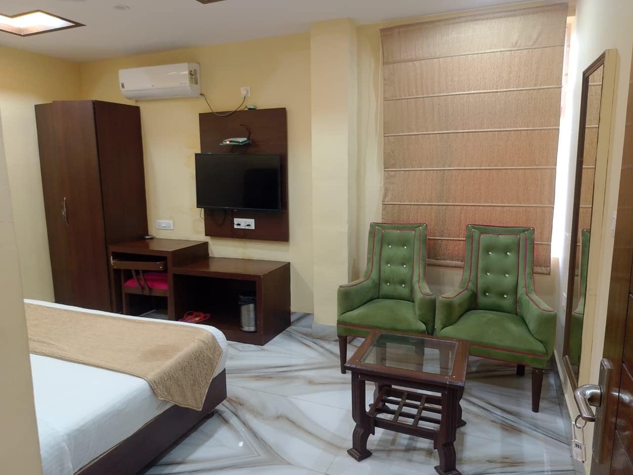 A hotel room with a bed, a desk, a television, two green armchairs, and a glass coffee table.