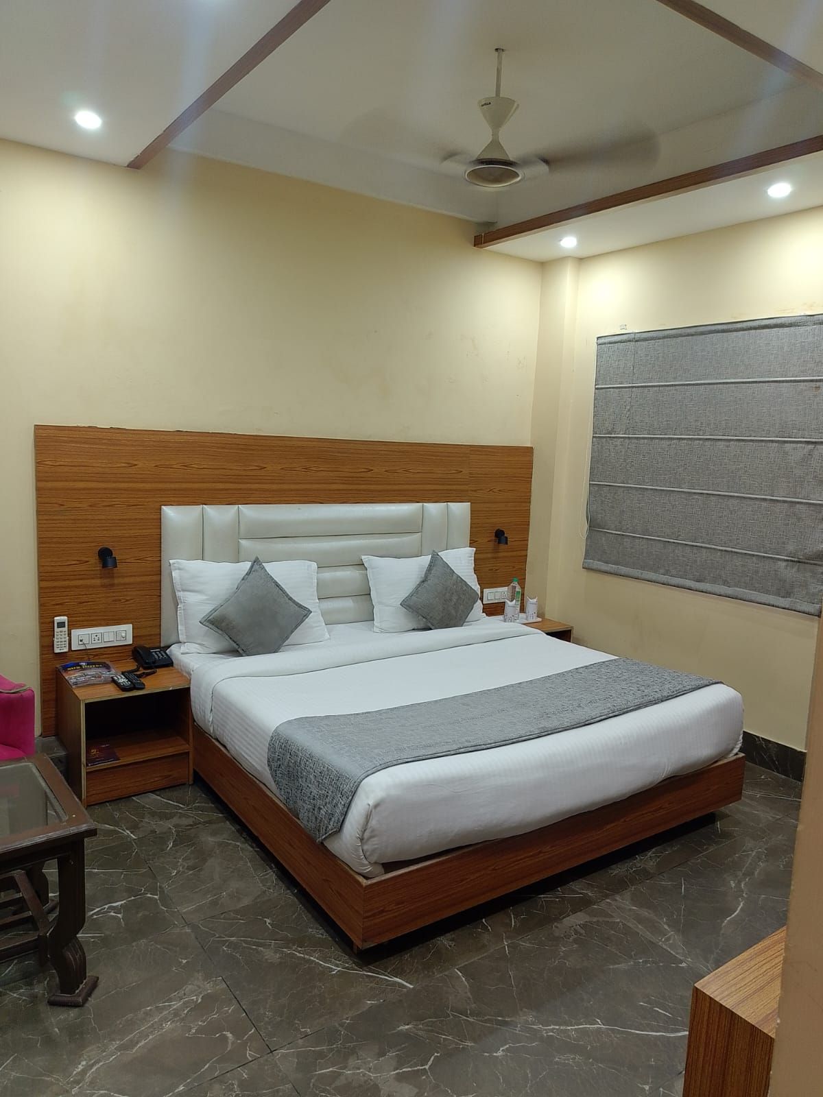 A hotel room with a bed, bedside table, nightstand, and a window with blinds.