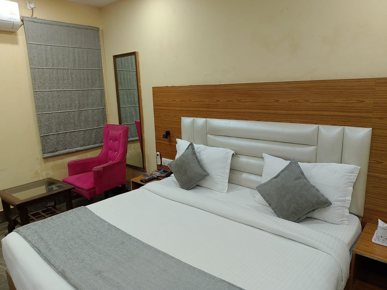 Una habitación de hotel con cama, una silla, una mesita de noche, un espejo y una ventana con cortinas.