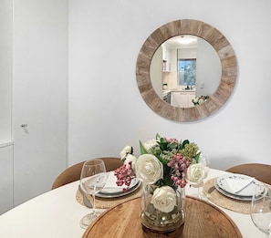 A dining table with a mirror above it, a vase with white flowers, and a view of a kitchen and a window. - Style & Bright 2BR with leafy balcony & parking (Cammeray)