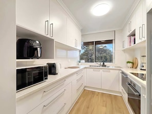 A compact kitchen with white cabinetry, a built-in microwave, a coffee maker, and a window providing a view of trees. - Style & Bright 2BR with leafy balcony & parking (Cammeray)