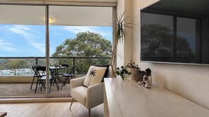 A modern living room with a sofa, a dining table with chairs, and a large window offering a view of trees. - Style & Bright 2BR with leafy balcony & parking (Cammeray)