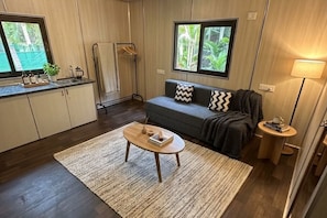 A compact living space with a sofa, a small coffee table, a side table, a window with a view of greenery, and a towel rack. - Coconut Grove Ecostay Stay with Pool Cinema & BBQ (Halaguru)