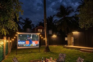 A backyard with a large screen displaying a cricket match, outdoor seating, and a house in the background.