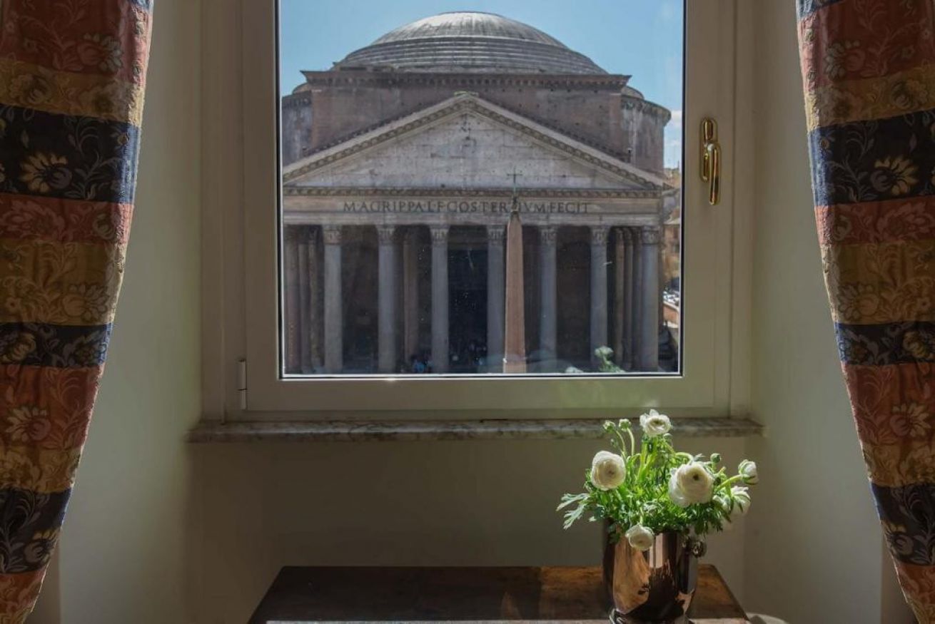 A window with a view of a classical building with columns and a dome, a vase with flowers on a table below.