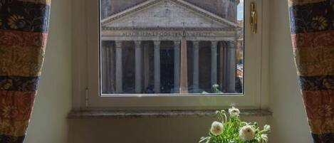 A window with a view of a classical building with columns and a dome, a vase with flowers on a table below.