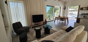 Ein modernes Wohnzimmer mit einem Flachbildfernseher, einer Couch, einem Essbereich mit Tisch und Stühlen und Blick auf einen Pool durch große Fenster.